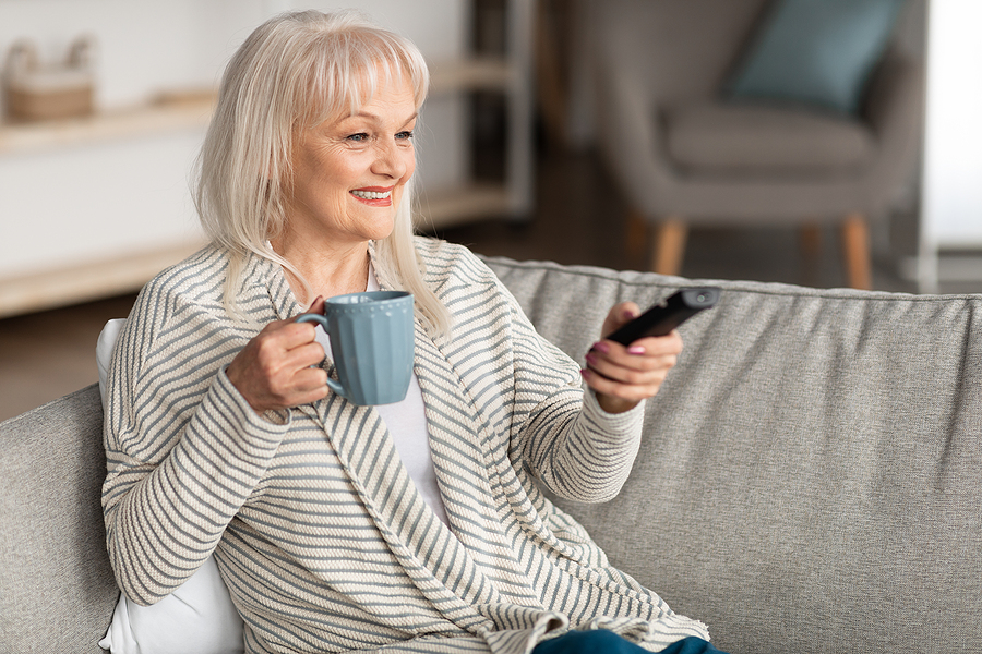 An older woman holds a remote and a cup of coffee.
