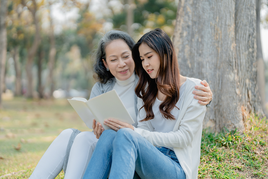 Grown daughter and adult grandmother sit under a tree reading