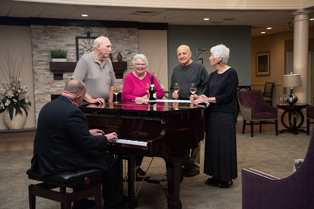 A group of friends gathers around a piano with drinks in hand.